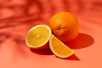 On the coral surface, next to two neatly sliced slices, there is a whole orange. Sunlight casts soft shadows, emphasizing the freshness and bright color of the fruit.