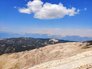 Panoramic mountain landscape with rocky hills, forested valleys and bright blue sky with a large white cloud. High-altitude nature view of wilderness terrain and distant mountain ridges.