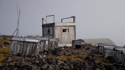 Abandoned wooden outpost with weathered cabins and scattered sheds on a rocky, mossy tundra, shrouded in fog and overcast light, evoking a bleak, remote, and desolate landscape. - Powered by Adobe