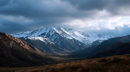 Majestic snow capped mountain range and valley illuminated by golden hour light beneath dramatic stormy clouds