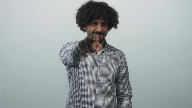 Man with curly hair pointing finger at camera in light gray studio wearing checked shirt and slight smile; confidence.