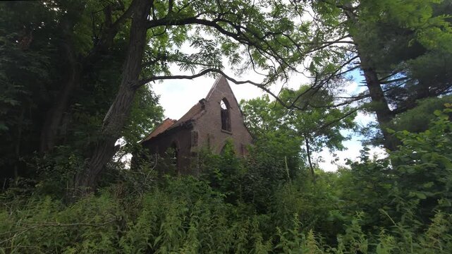 Exterior view of a neglected, abandoned brick church or chapel ruin, showing the rounded apse and an arched window, surrounded by a dense, overgrown forest and trees