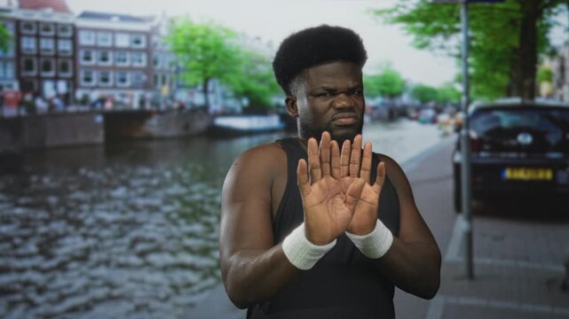 Man holding hands up palm out on a city street by an amsterdam canal, wearing black tank top and white wristbands, face scowl; boundary refusal annoyance.