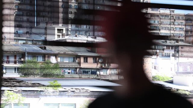Silhouette of a person looking through window blinds at old residential buildings. View of dense urban architecture and apartment blocks in a city from the inside.

