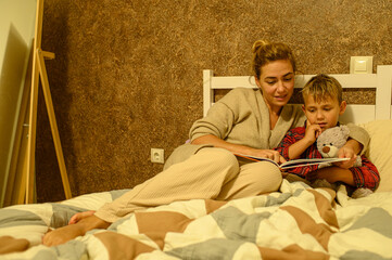 Mother and son at home preparing for bedtime, putting on pajamas, reading a book, mother kissing her child and tucking him into bed. Family bonding, love, bedtime routine.