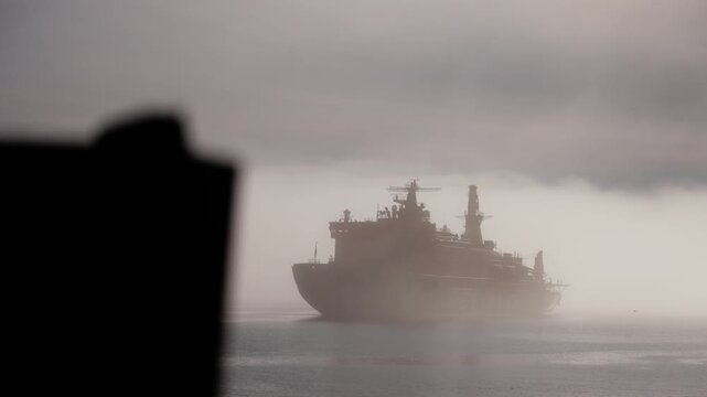 Large seagoing vessel emerges through dense fog on a calm, gray ocean, its silhouette and superstructure looming beneath an overcast sky, creating an atmospheric maritime scene.