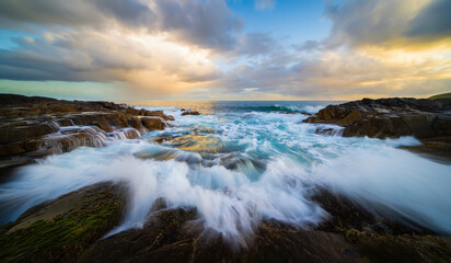 A rocky coastal seascape with churning waves crashing onto tiered rock formations. Dynamic motion of the turbulent turquoise water under a dramatic evening sky
