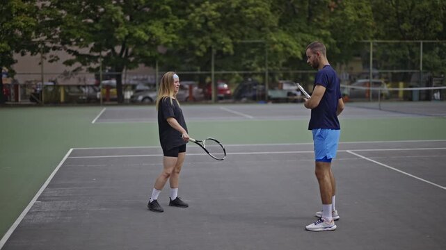 Two friends, a man and a woman, are on a green tennis court, rackets in hand. They appear to be discussing strategy or taking a break. A perfect summer day for an outdoor sport.