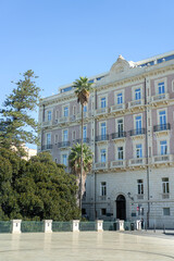 Obraz premium Elegant historic building with classical facade and balconies, surrounded by palm trees and greenery in Syracuse, Sicily. Southern Italian urban architecture under a clear blue sky