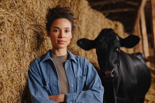 Young female farmer poses beside a cow in a dairy barn filled with hay