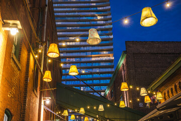 Fototapeta premium Decorative hanging lamps and string lights illuminating a narrow alley between historic brick buildings in the Distillery District of Toronto at night.
