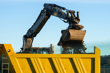 Excavator unloading soil into large yellow dump truck bed at construction site on clear sunny day