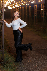 A young woman is striking a confident pose in a picturesque outdoor setting, illuminated by warm lights during the night