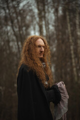 Man with long curly hair in a dark coat stands in a misty forest during autumn