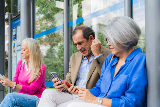 Group of diverse senior friends sitting at a bus stop, engaging with technology