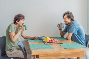 Older woman having breakfast at the table with her adult son, enjoying a warm family moment at home. Morning routine, healthy lifestyle and togetherness concept, representing connection, care and