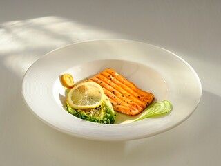 A piece of grilled salmon is artfully arranged with lettuce and a lemon slice on a white plate. Light from the window adds a warm glow to the presentation