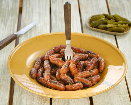 A yellow plate overflows with small, browned sausages, speared by a fork. Pickles rest nearby on another plate, all on a textured wooden surface with a knife