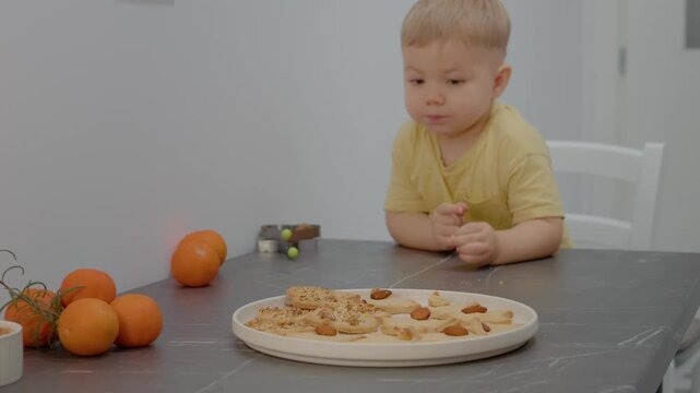 A young boy gazes longingly at a plate of cookies arranged neatly on a kitchen table. He sits quietly, surrounded by fruit, eager for a treat.