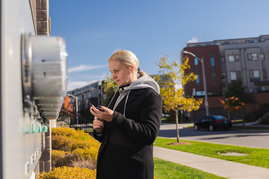 Middle-aged woman checking electricity meters outside a residential building and entering usage data on her smartphone during a sunny autumn day