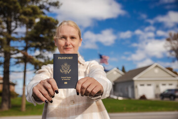 Woman standing outdoors and holding a United States passport toward the camera, with houses, trees and a blue sky in the background.