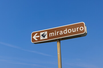 Close-up of a brown 'Miradouro' (viewpoint) directional sign in Portugal, with a white arrow and binocular icon against a clear blue sky.