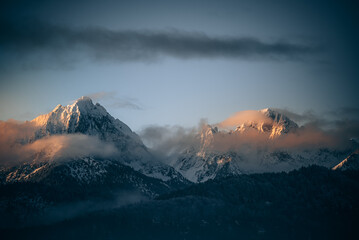 View of snow-capped mountains piercing through the swirling clouds as the sun kisses their peaks with golden light, Schwangau, Bavaria, Germany.