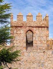 Close-up of an &Aacute;vila wall tower with cylindrical stone blocks, notched parapets, and an arched opening framed by reddish brickwork, with green foliage at left under a blue sky