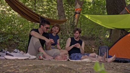 A cheerful group of young friends enjoys a lively card game while camping amidst lush green trees. They are relaxing outdoors, sharing laughter and good times, making memories together in nature.