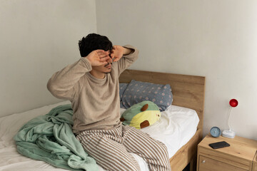 Man sitting on the bed upon waking up, stretching and covering his eyes as he starts his morning with both tiredness and calm. Everyday scene in his bedroom
