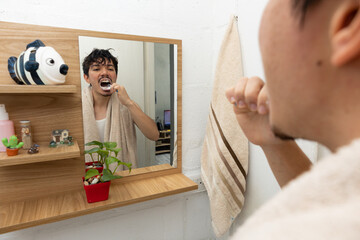 Man in the bathroom with a towel around his neck, brushing his teeth from a rear perspective that shows his reflection in the mirror. Morning hygiene routine at home