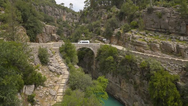 White van and yellow bus traveling on an ancient stone bridge over a turquoise river in Koprulu canyon, Turkey