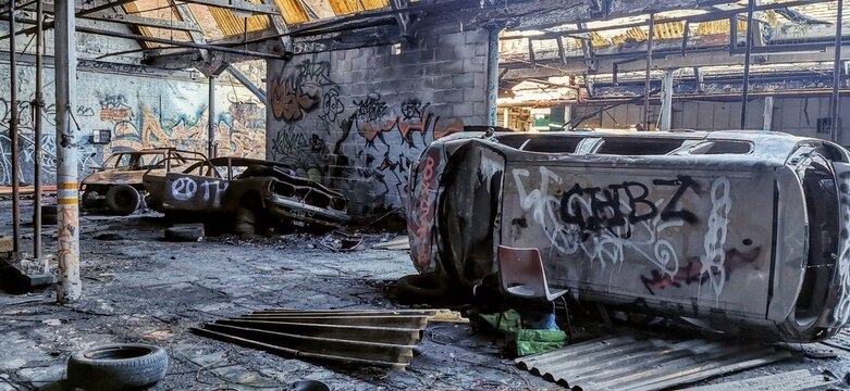 Graffiti-covered overturned cars, fire damaged collapsed roof warehouse. Twisted burnt out shell, metal scattered debris. Spray paint vandalized vehicle. Auto scrap-yard accident wreckage aftermath.