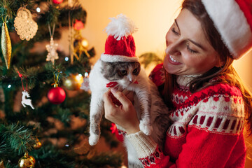 Celebrating Christmas with cat by New Year tree at home. Woman holding pet with Santa's hat on...