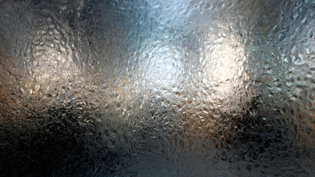 Textured glass surface covered in water droplets, creating an abstract pattern with shimmering light reflections and varying translucency. - Powered by Adobe