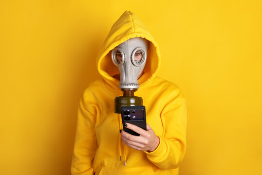 Individual in yellow hoodie and gas mask holding smartphone against vibrant yellow background, symbolizing technology, safety, and modern challenges. - Powered by Adobe