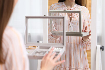 Woman with jewelry box choosing accessories near mirror indoors