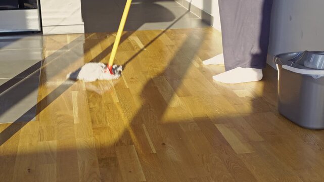 A person efficiently mops a gleaming wooden floor, reflecting bright sunlight, within a clean modern kitchen. This diligent household chore maintains a fresh and hygienic home environment.
