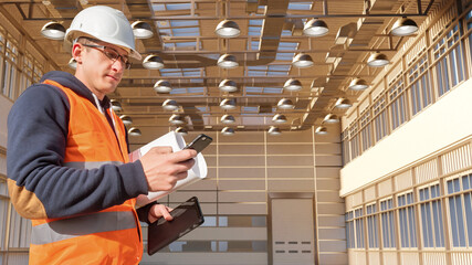 Construction site supervision involves checking building plans and coordinating tasks, as the inspector reviews documents and communicates updates using a mobile device. © Grispb