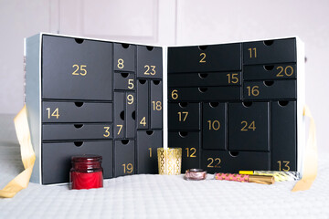 Black numbered advent calendar box with small beauty gifts and candles in front, festive holiday countdown set on a soft white surface.
