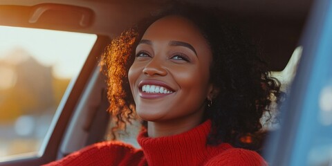 A woman with curly hair is smiling in a car. She is wearing a red sweater. The car is parked on the side of the road