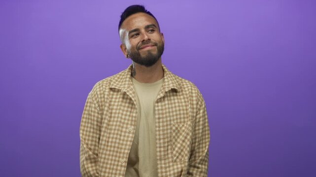 Young hispanic man in beige checkered shirt smiling while looking up at camera in studio with purple wall; contentment quiet reflection.
