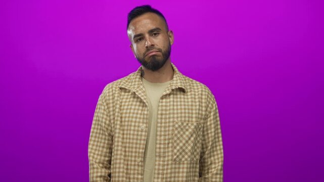 Young hispanic man smiling with visible face and beard wearing plaid shirt in studio against magenta purple backdrop; confidence.