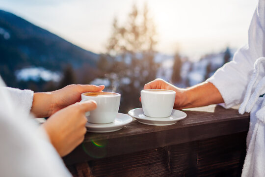 Close up of hot coffee. Loving couple drink beverages on hotel balcony enjoying winter mountains view wearing white robes. - Powered by Adobe