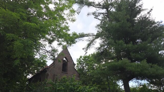 Exterior view of a neglected, abandoned brick church or chapel ruin, showing the rounded apse and an arched window, surrounded by a dense, overgrown forest and trees