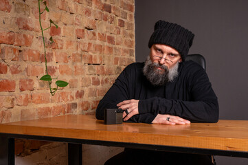 Portrait of a serious bearded hipster man in glasses and beanie sitting at a table with a smartphone. Creative freelancer in a loft office against a brick wall background