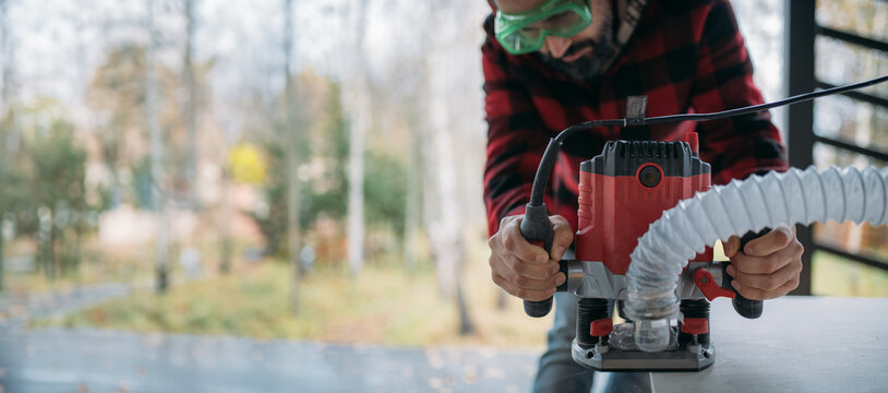 A young man is working with a milling cutter on the veranda of a house. The carpenter chamfers the plywood using a milling machine.