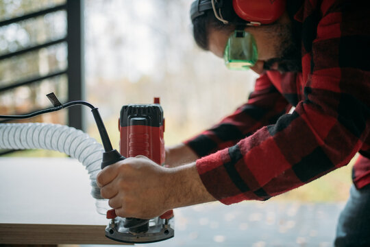 A young man is working with a milling cutter on the veranda of a house. The carpenter chamfers the plywood using a milling machine.