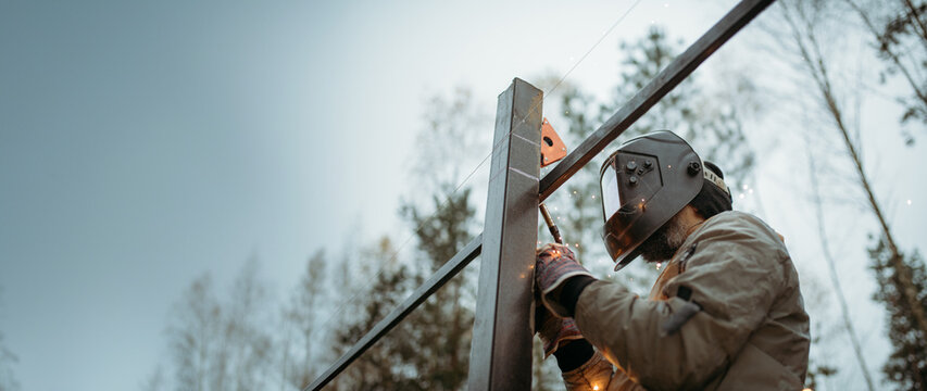 A young man is working as a gas welder on the street. A man wearing a protective mask is welding iron rungs to pillars. The worker, holding a welding machine, sparks fly around him.