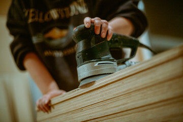 A young woman grinds plywood in a house under construction. Close-up of a woman's hands holding a grinder. A woman works with a manual electric grinder